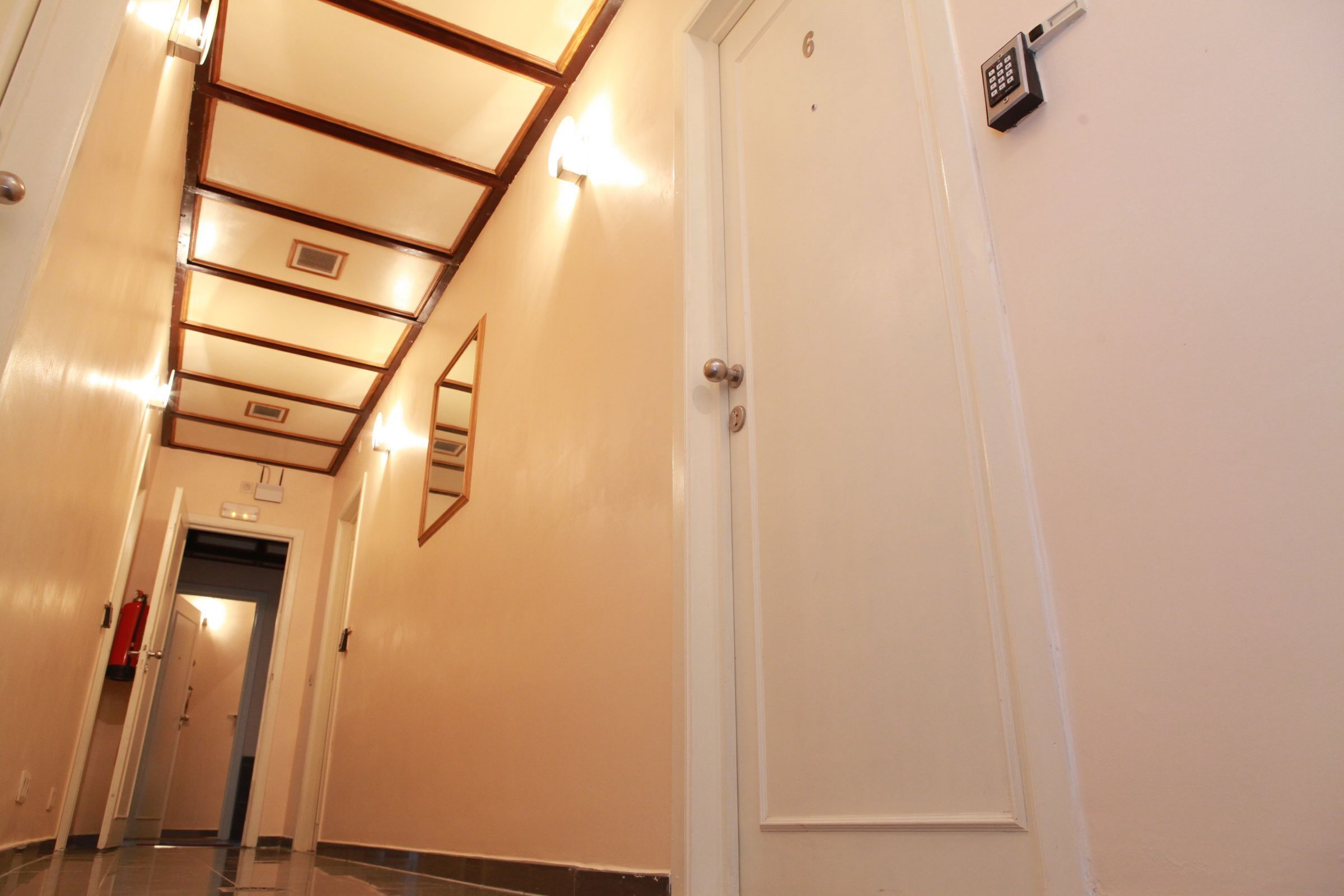 Un couloir d’hôtel avec une porte blanche, un extincteur et un clavier de sécurité fixé au mur.
