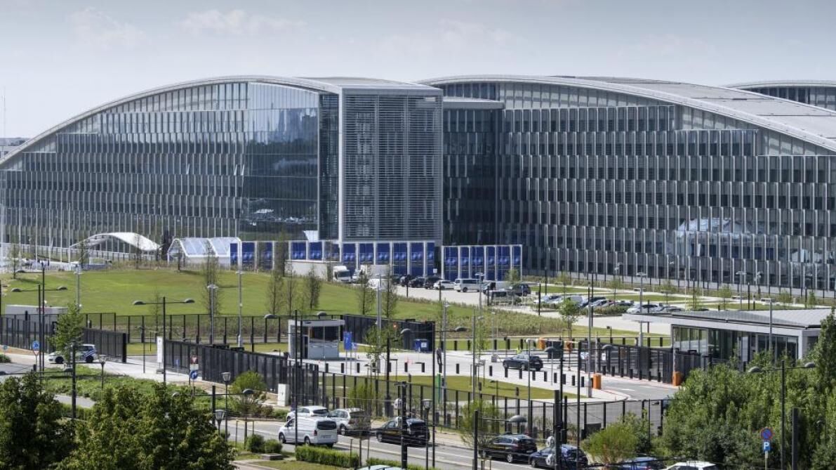 A modern building with a curved glass facade, surrounded by a parking area and greenery.