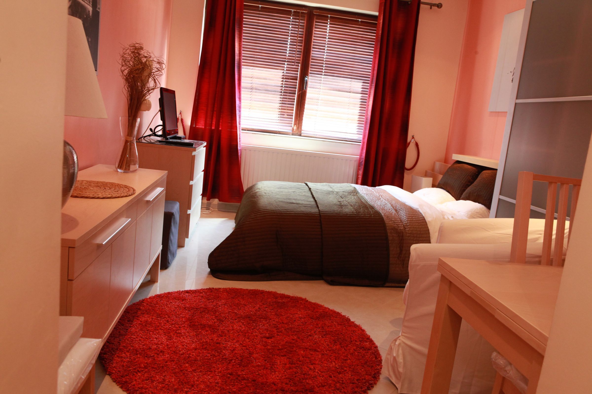 Une chambre à coucher avec un lit, un bureau, une chaise, une fenêtre avec des rideaux et un tapis rouge.