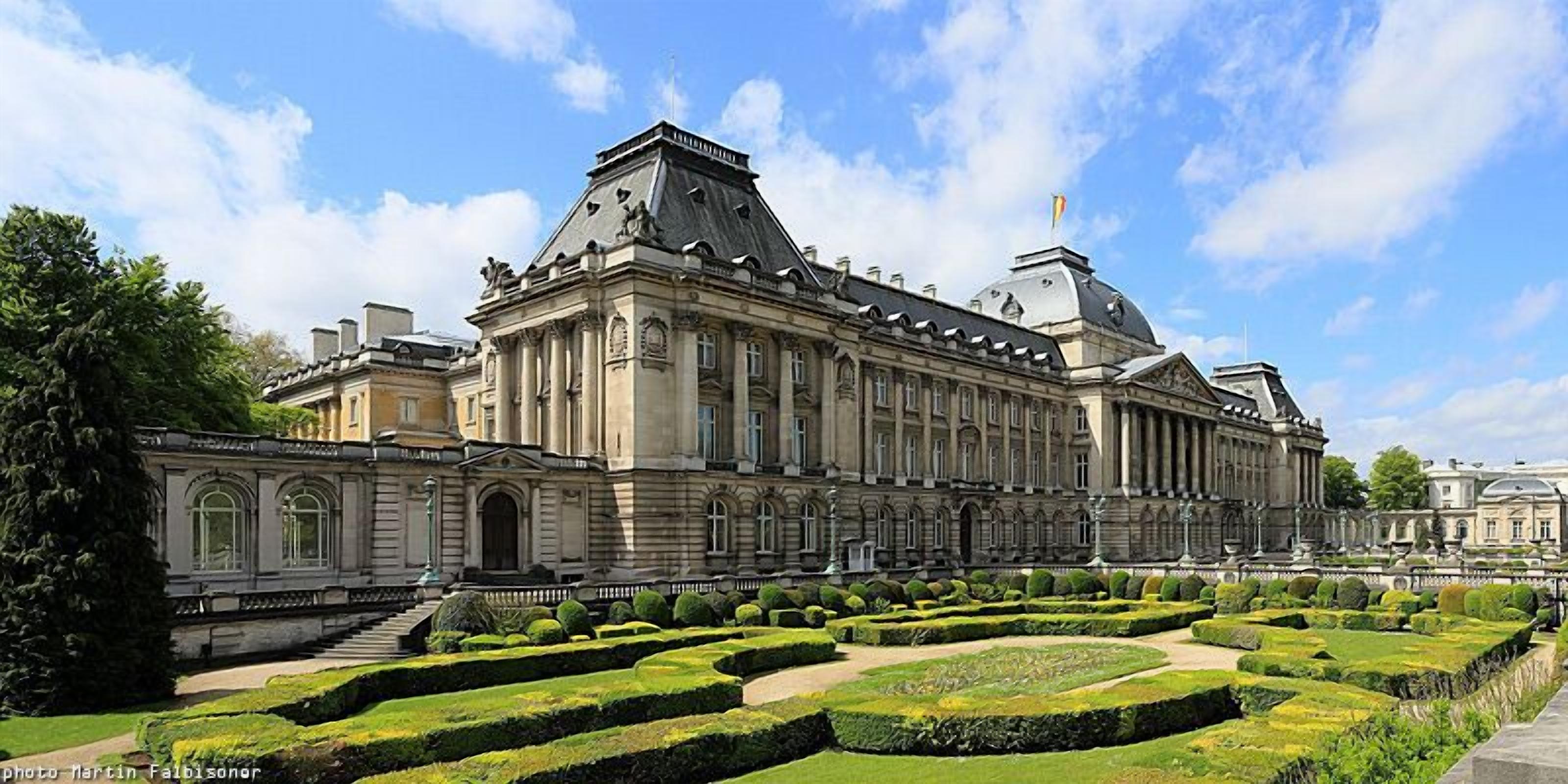 A large, historic building with a symmetrical facade, formal garden with geometric hedges, and a central staircase leading to the entrance.