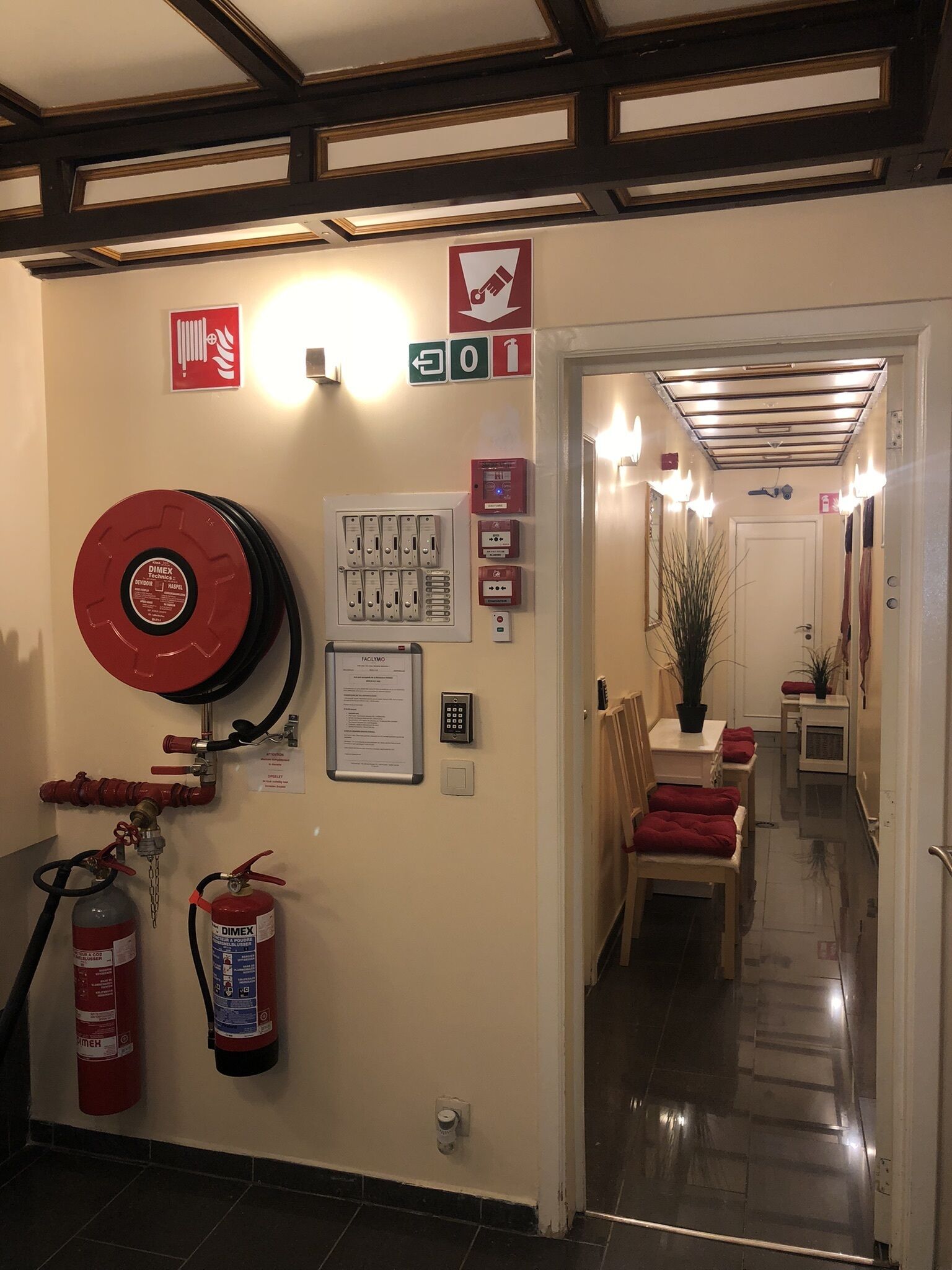 A hallway with a fire extinguisher, emergency exit sign, and a fire alarm panel.