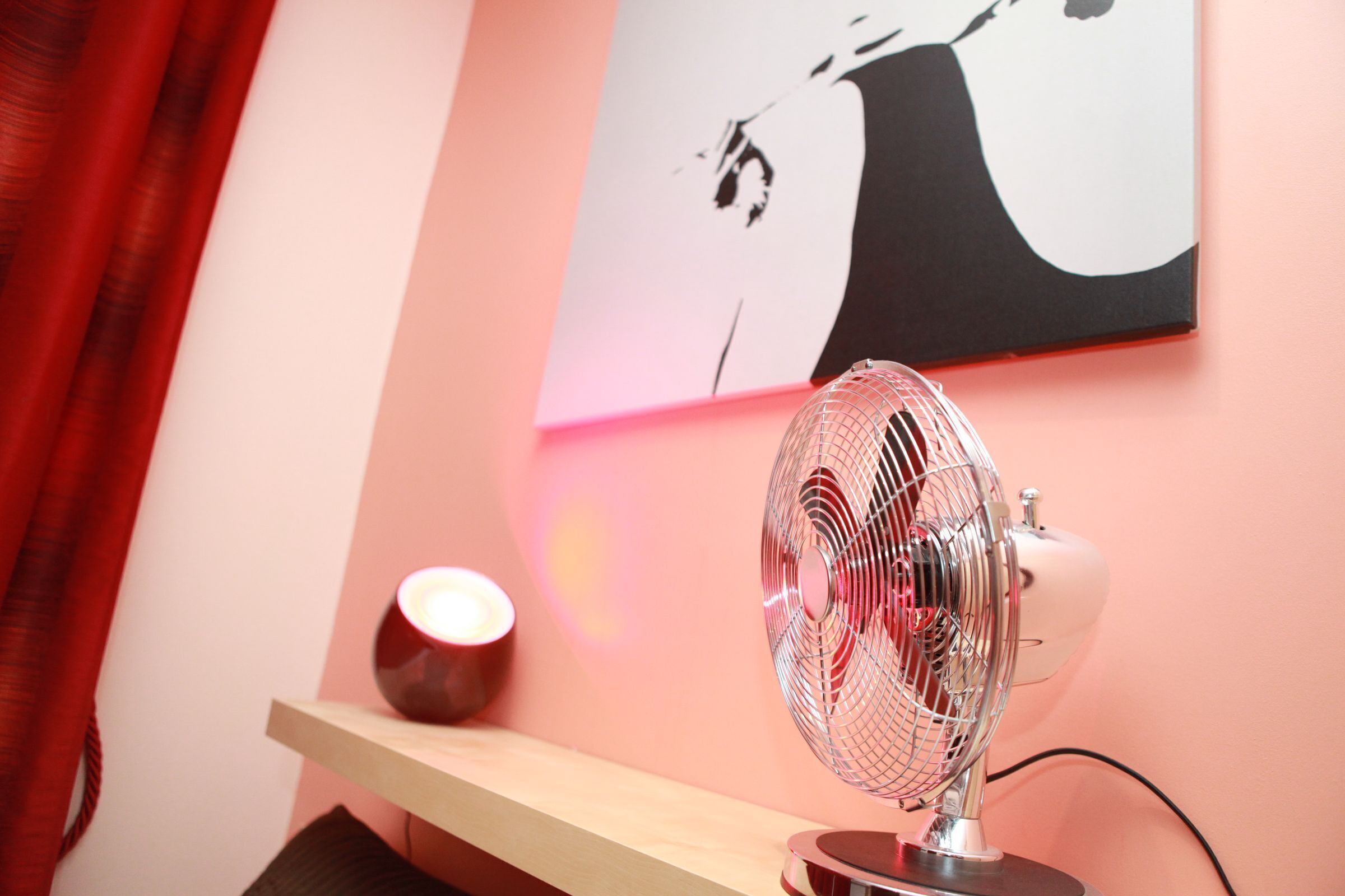 Una habitación con un ventilador de pie, una lámpara roja y un cuadro colgado en la pared.