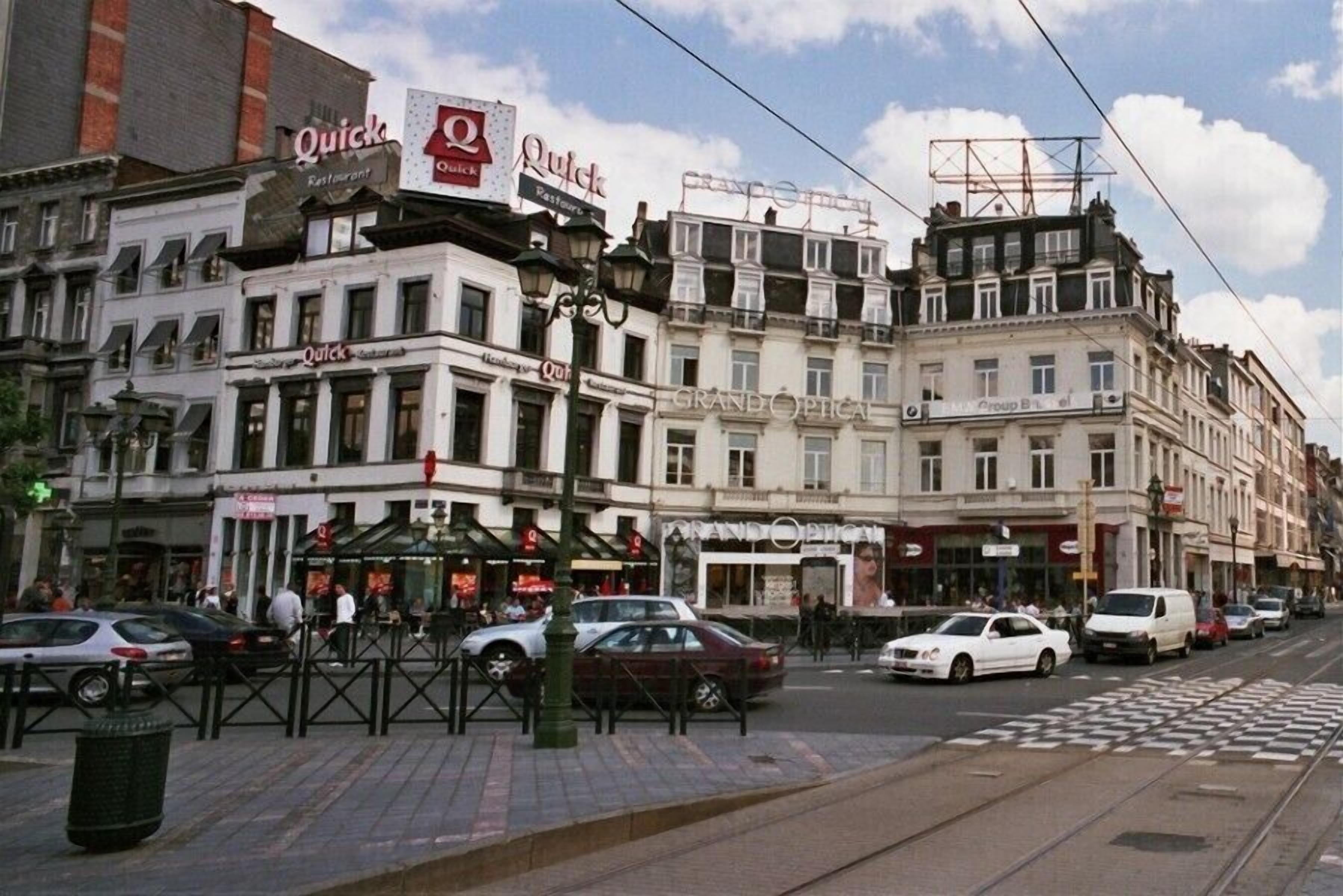 A street with multiple buildings, including a restaurant named 'Quick' and a pharmacy named 'GRANDOPTICAL'.