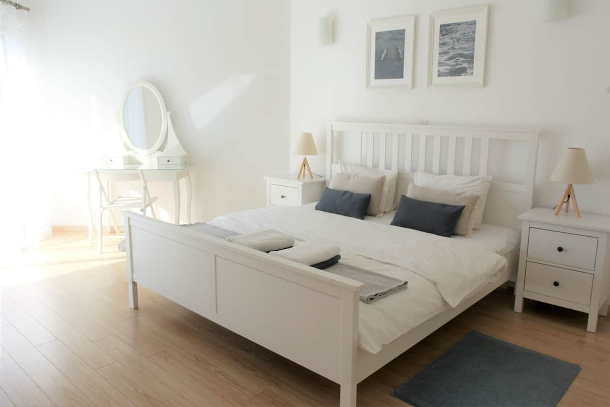 A white bedroom with a bed, bedside tables, a mirror, and two framed pictures on the wall.