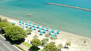An aerial view of a beach with rows of blue umbrellas and beach chairs, a boat in the water, and a rocky breakwater.