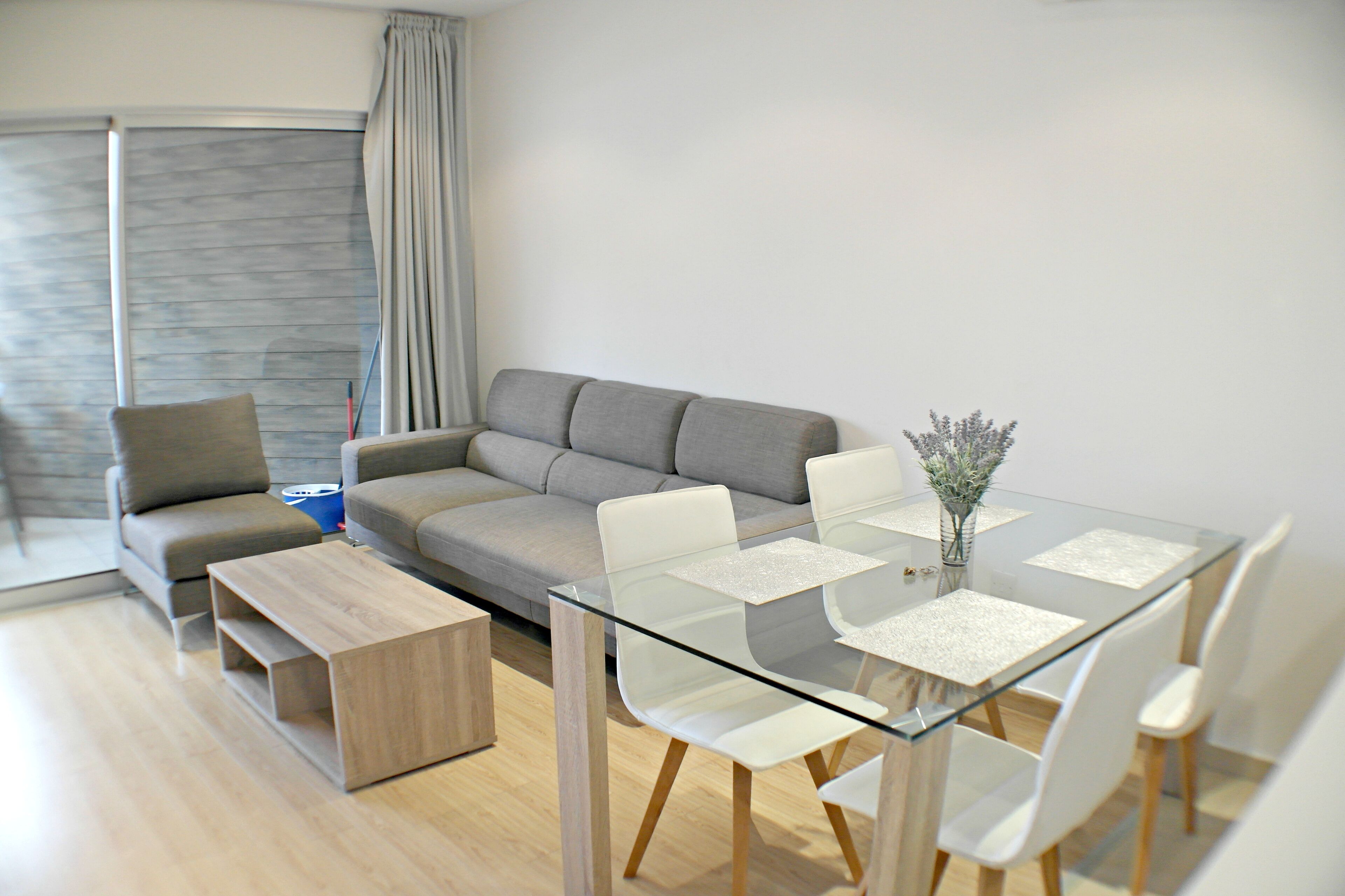 A modern living room with a glass dining table, white chairs, a grey sofa, and a wooden coffee table.