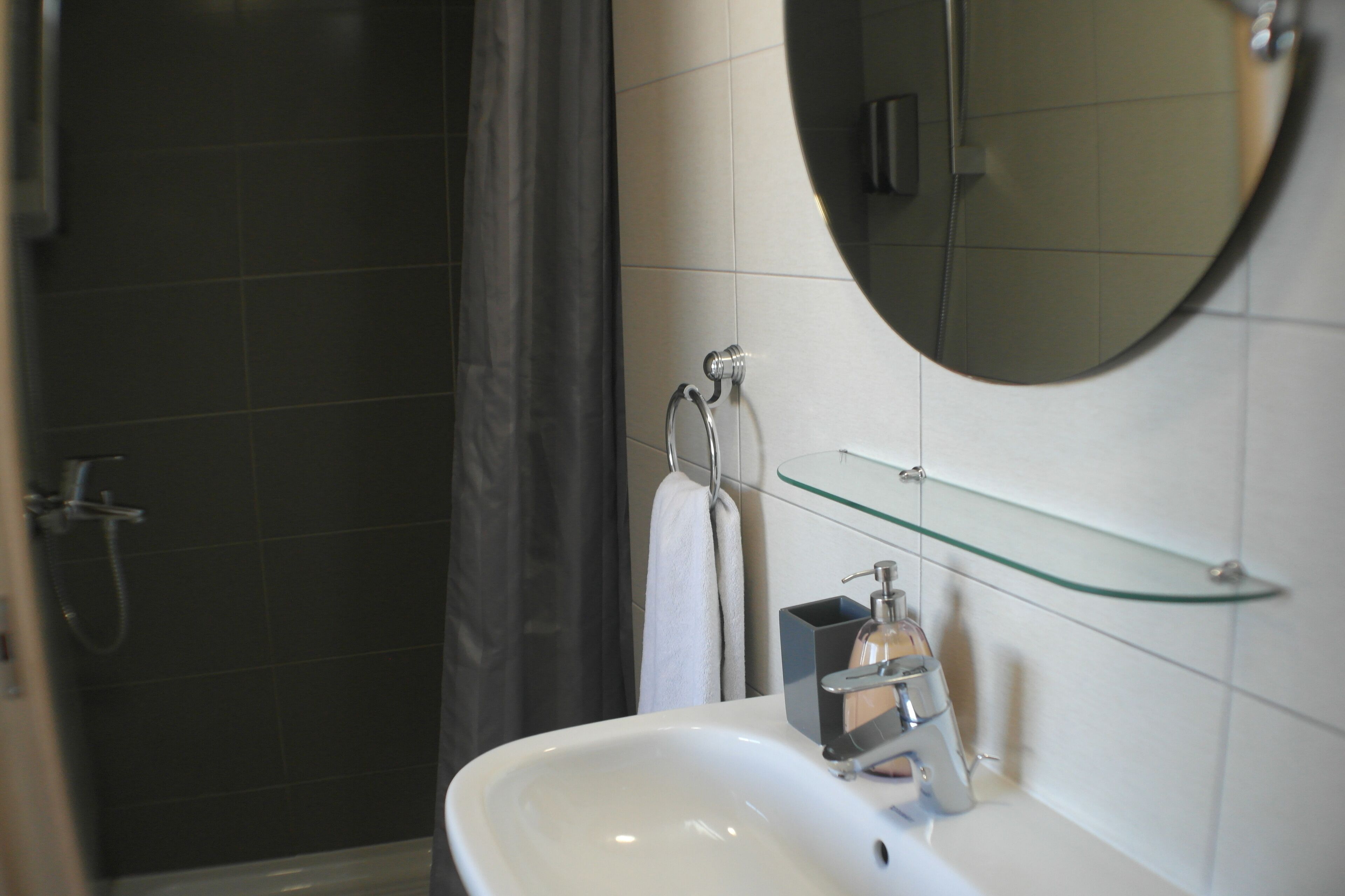 A bathroom with a white sink, a glass shelf, a round mirror, and a shower area.