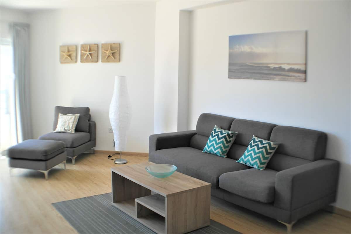 A modern living room with a grey sofa, a grey armchair, a glass coffee table, and a large vase. There are three starfish wall art pieces and a painting of a beach scene.