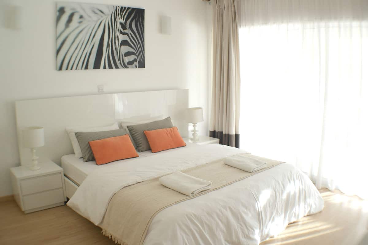 A neatly made bed with a white headboard, two pillows, and a beige throw blanket. There are two bedside tables with lamps on either side of the bed.
