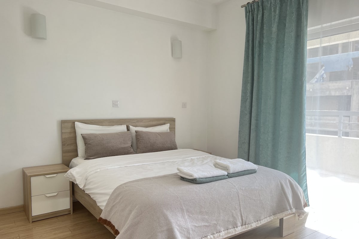 A modern bedroom with a wooden bed frame, white bedding, and a view of the outside through a window with a green curtain.