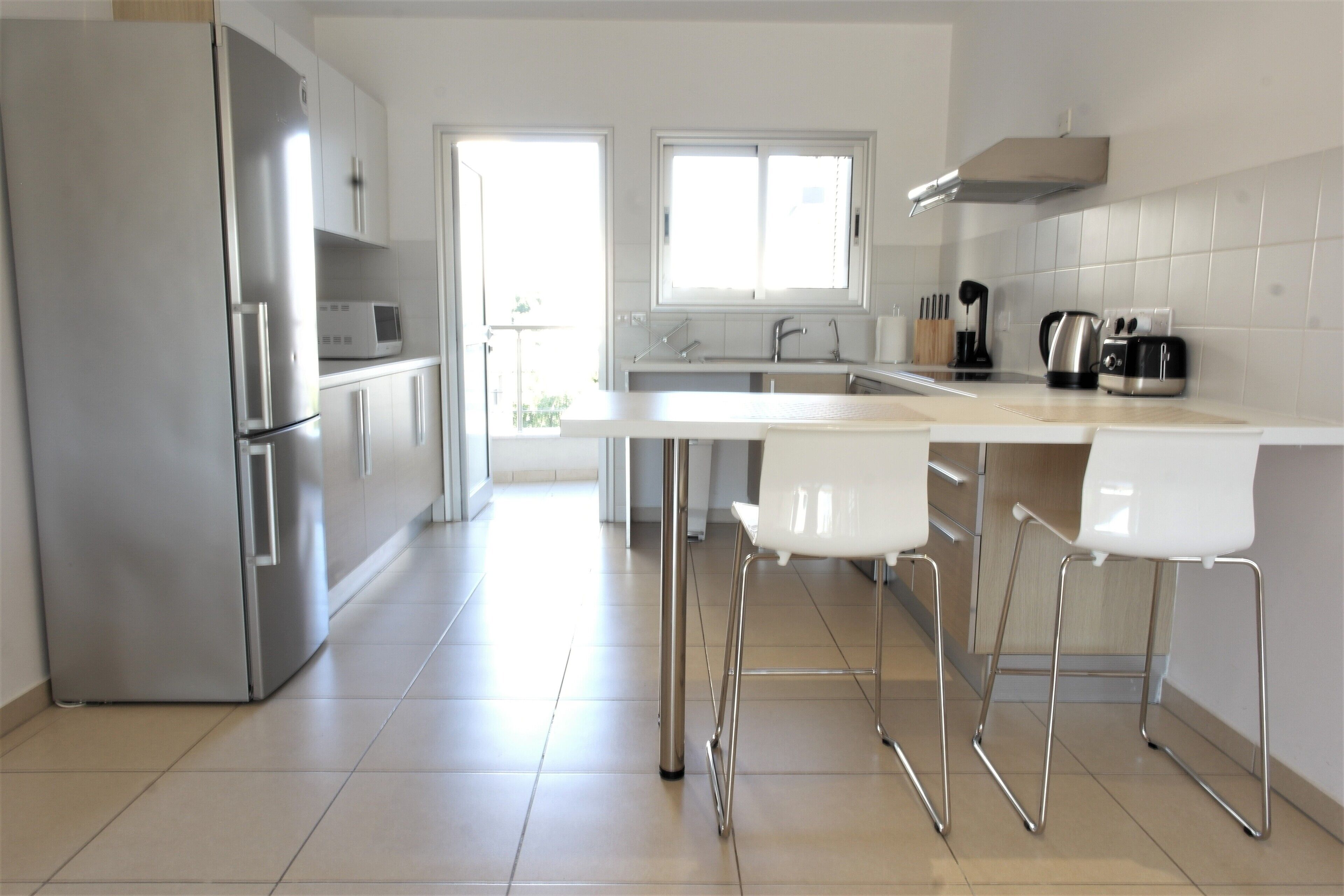 A modern kitchen with a stainless steel refrigerator, white bar stools, a microwave, and a window providing natural light.