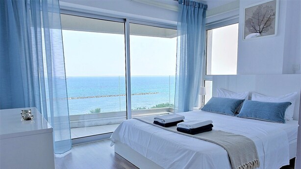 A modern bedroom with a large bed, a bedside table, and a view of the sea through floor-to-ceiling windows.