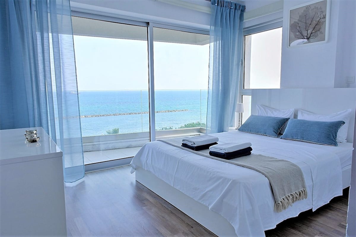 A modern bedroom with a large bed, a bedside table, and a view of the sea through floor-to-ceiling windows.