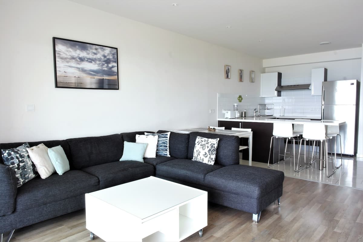 A modern living room with a sectional sofa, a white coffee table, and a kitchen area with white appliances and bar stools.