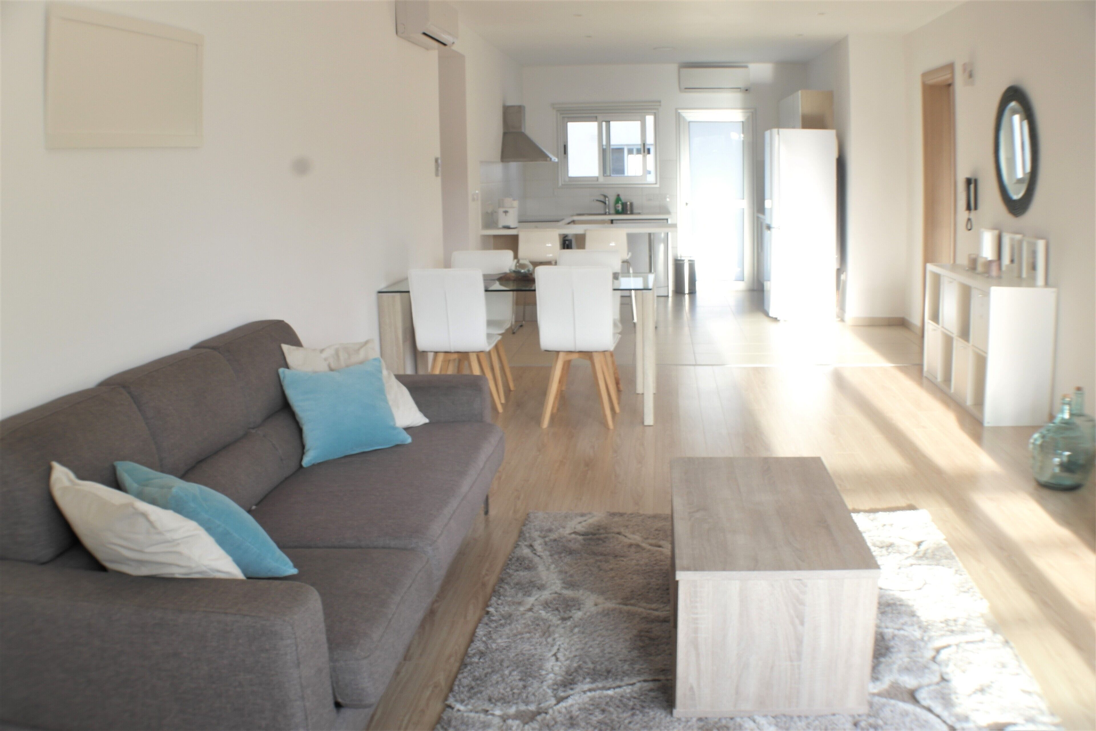 A modern living room with a grey sofa, a coffee table, and a dining area with chairs and a kitchen in the background.