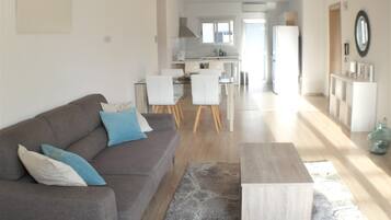 A modern living room with a grey sofa, a coffee table, and a dining area with chairs and a kitchen in the background.