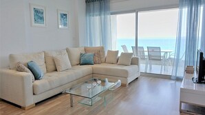 A modern living room with a beige sofa, glass coffee table, and a view of the sea through large windows.