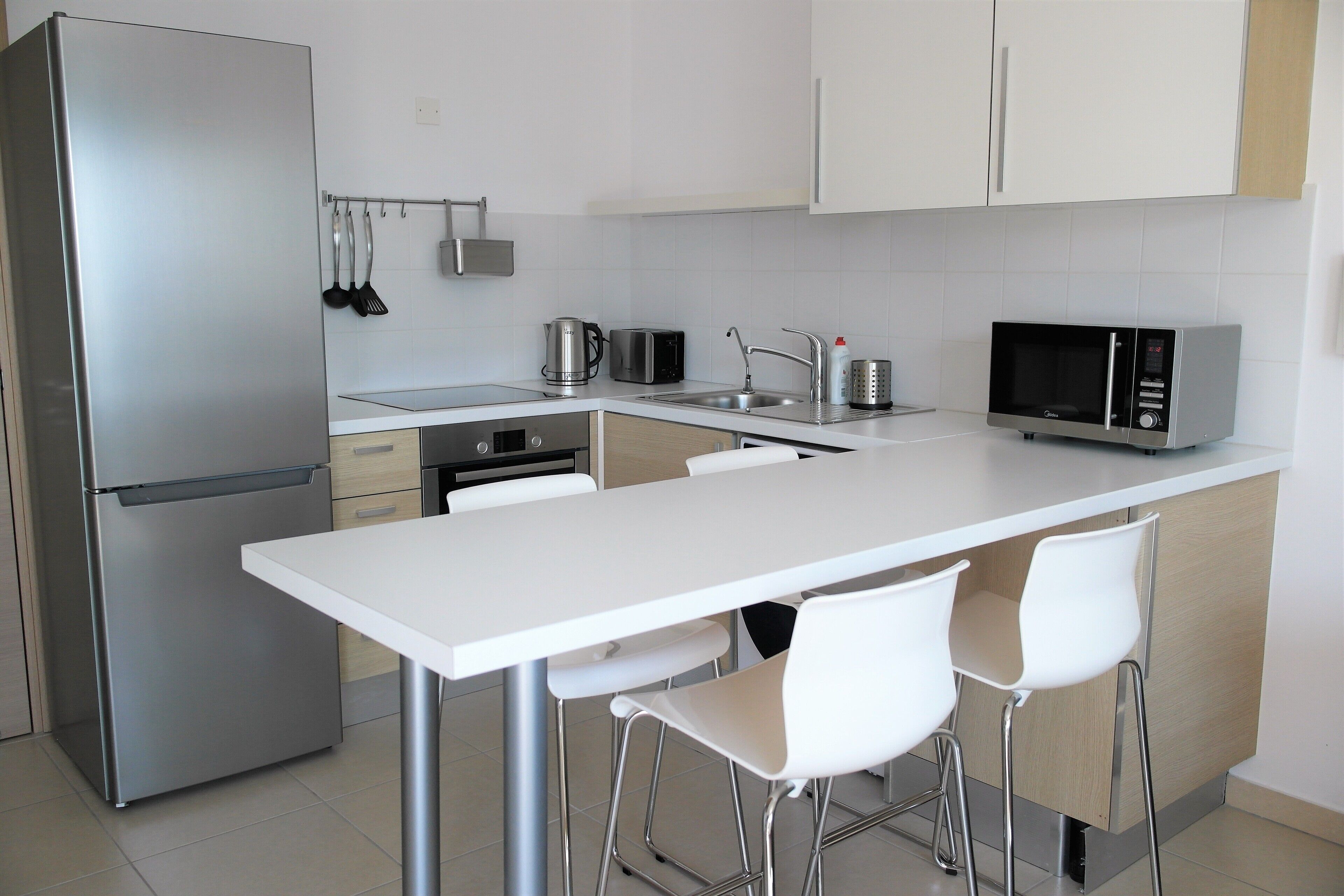 A modern kitchen with a stainless steel refrigerator, a microwave, a toaster, and a sink. There are two white bar stools and a white countertop.