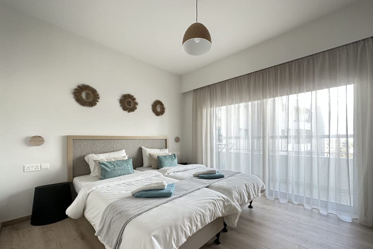 A modern bedroom with a large bed, a headboard, a bedside table, and a window with sheer curtains.