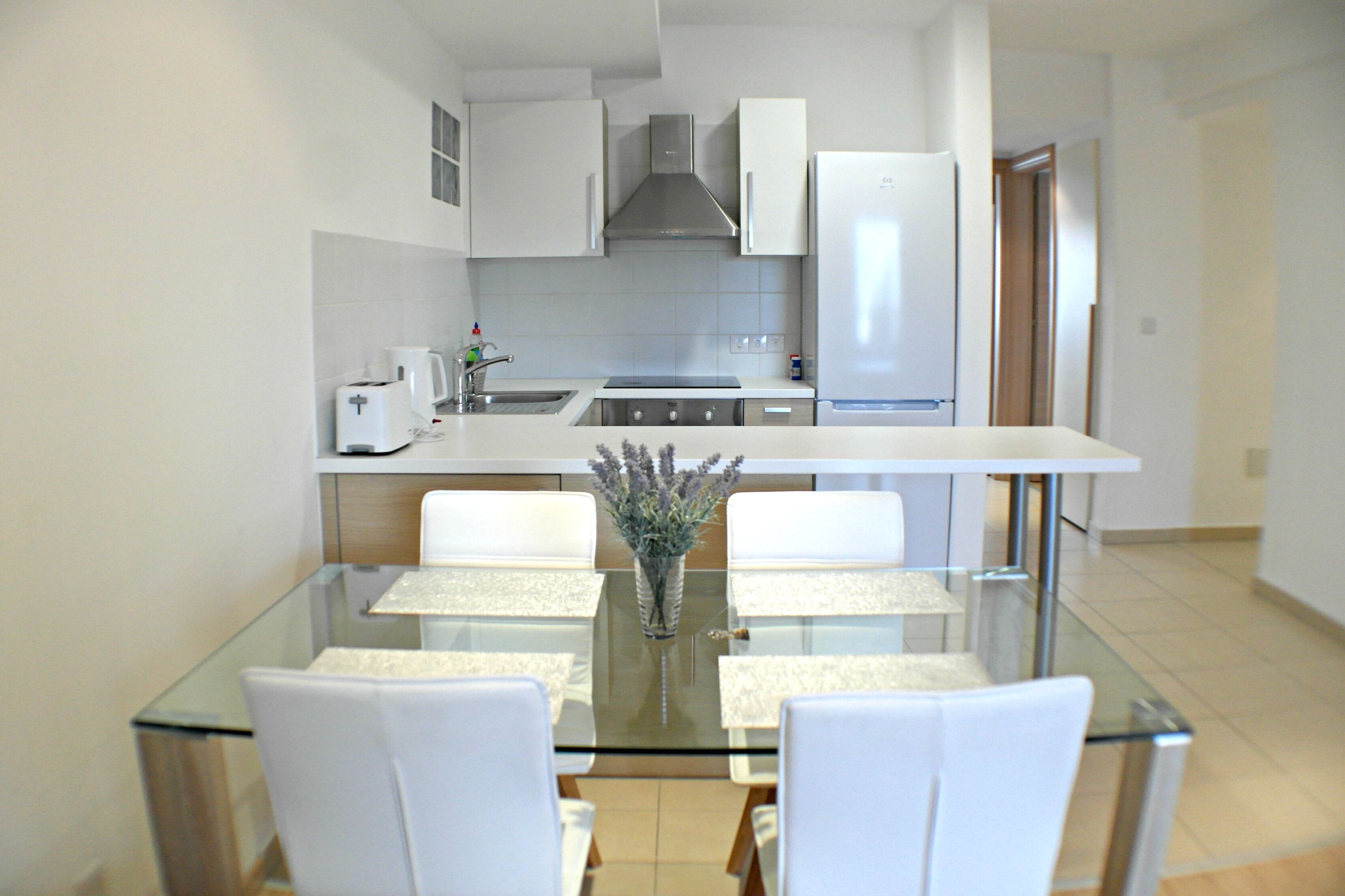 A modern kitchen with white cabinets, a stainless steel range hood, and a built-in oven. There is a dining area with a glass table and white chairs.