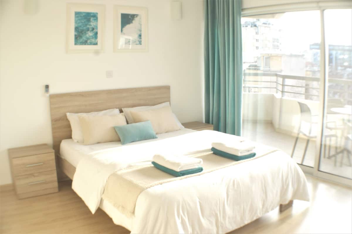 A hotel room with a large bed, a nightstand, a dresser, and a balcony with a view of the city.