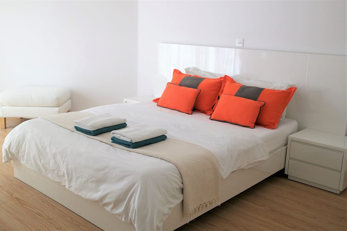 A neatly made bed with white bedding and orange pillows, a white nightstand with a drawer, and a white chair in a bright room with wooden flooring.