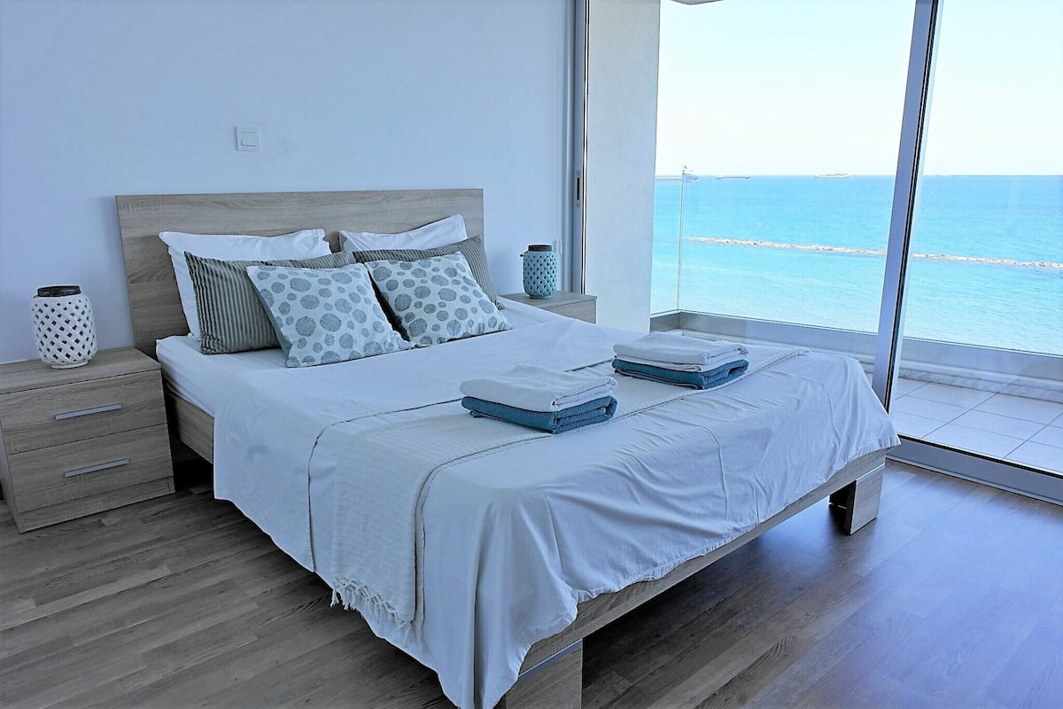 A modern bedroom with a large bed, wooden headboard, and a view of the sea through a large window.