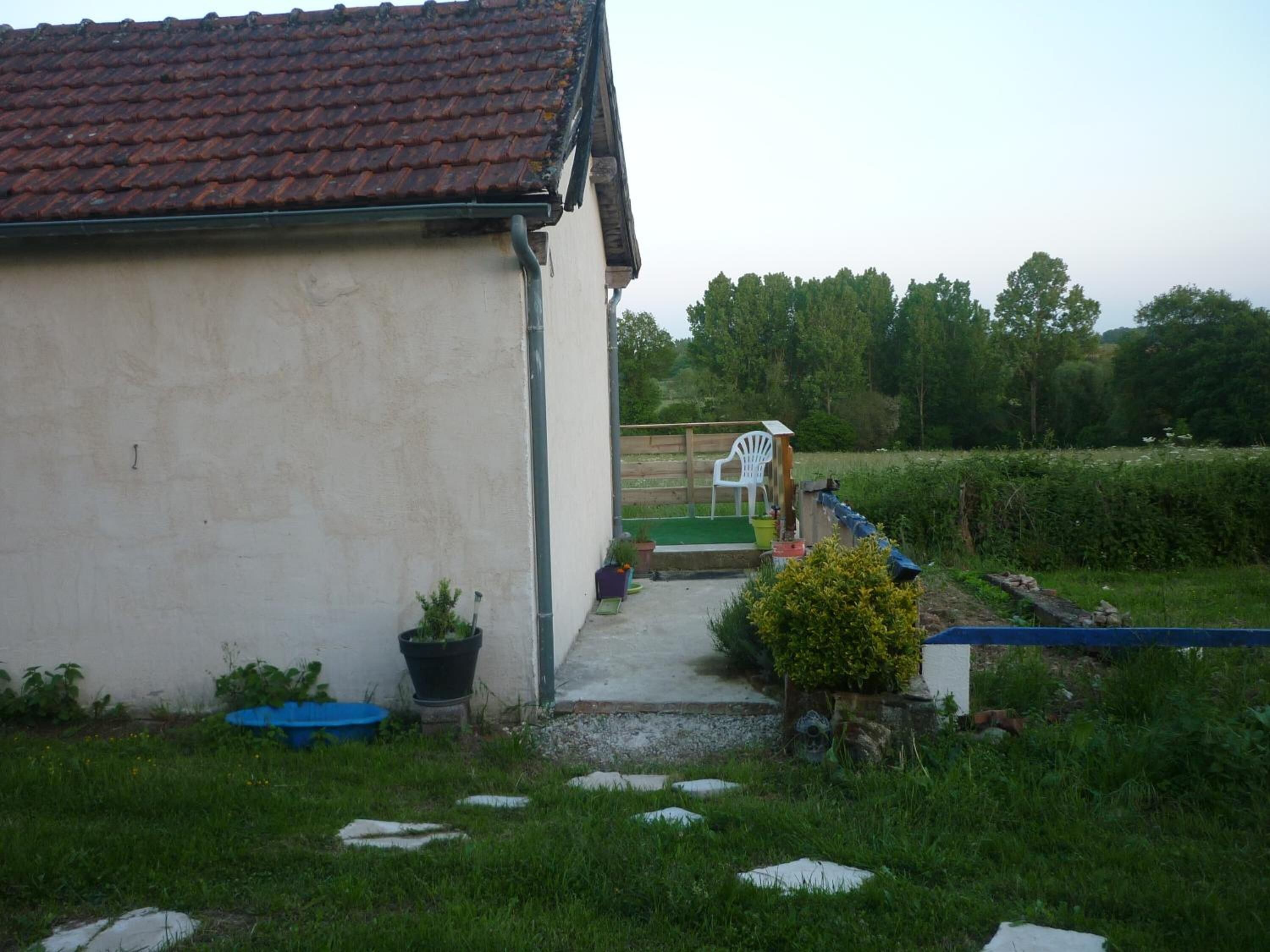 A small, single-story building with a tiled roof, a concrete pathway leading to a wooden deck, and a grassy yard with a few potted plants and a blue container.