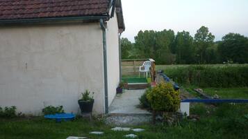 A small, single-story building with a tiled roof, a concrete pathway leading to a wooden deck, and a grassy yard with a few potted plants and a blue container.