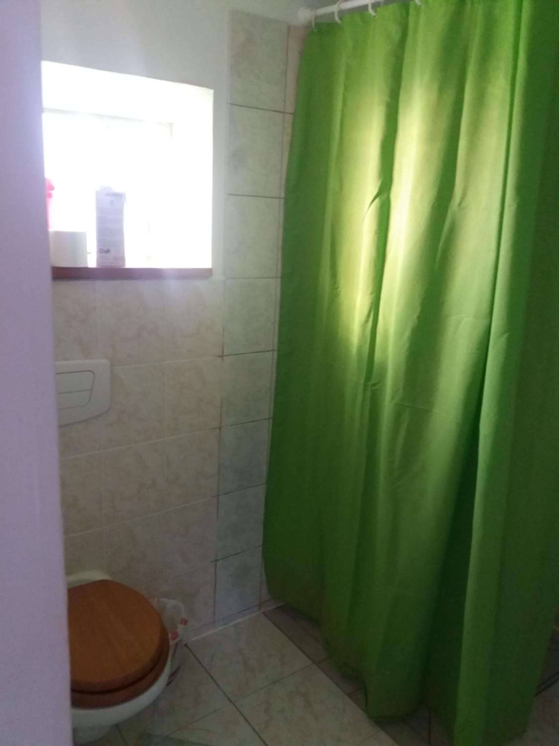 Un baño con cortina de ducha verde, un inodoro con asiento de madera y una ventana sobre el inodoro.