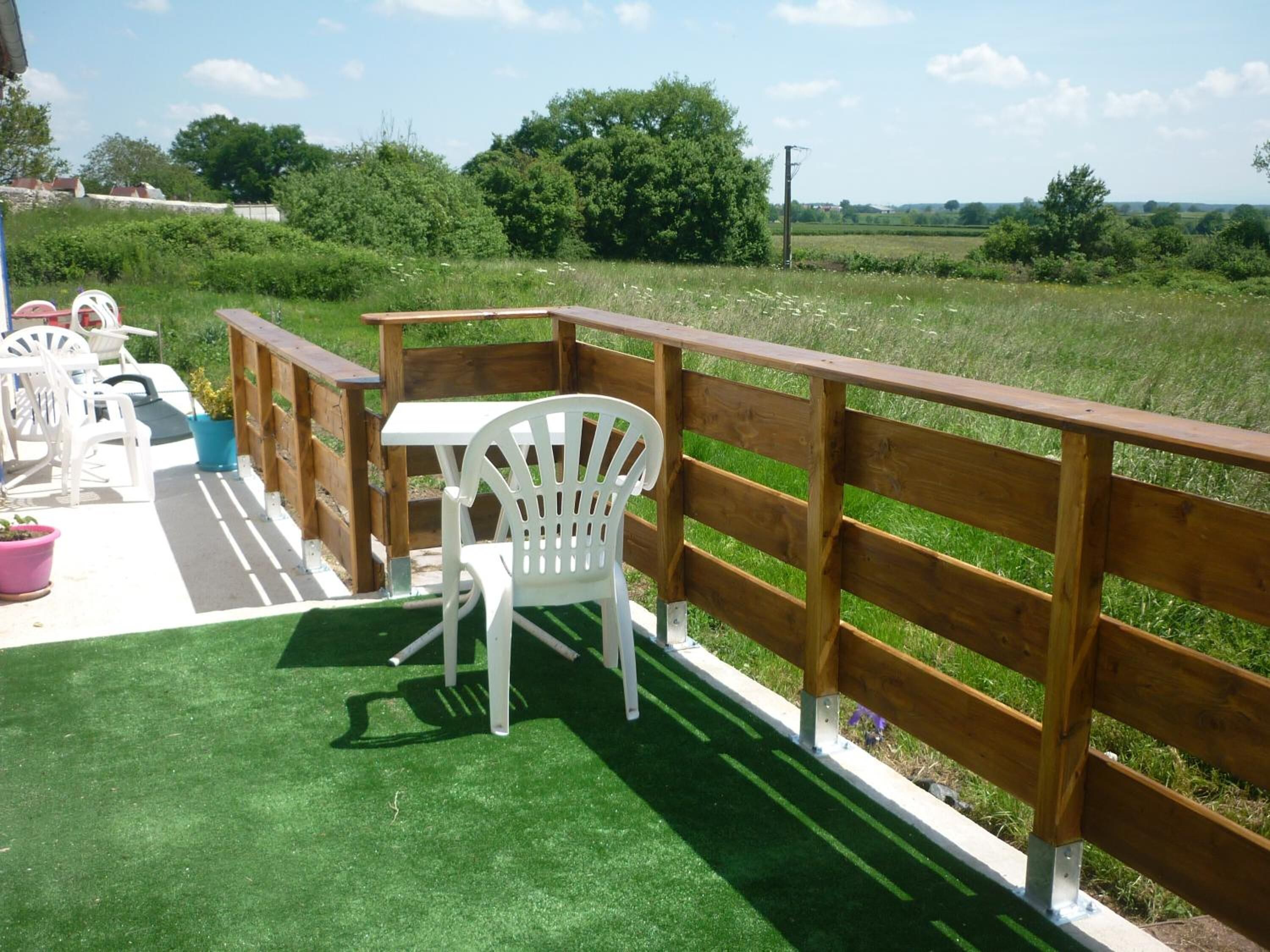 Een terras buiten met een houten terras, witte plastic stoelen en een tafel.