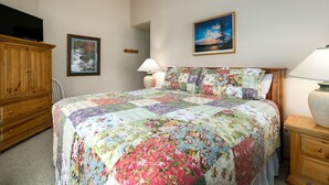 Un dormitorio con colcha acolchada de motivos florales, un aparador de madera, un televisor a la izquierda y un cuadro en la pared.