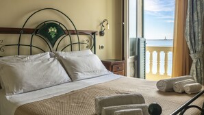 Uma cama com lençóis brancos, cabeceira com desenho floral e vista para o mar através de uma porta aberta.