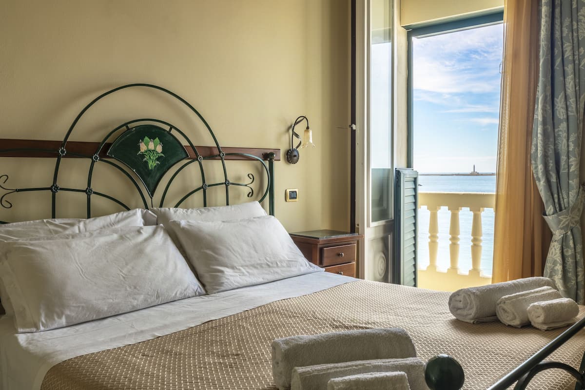 Un lit avec du linge de lit blanc, une tête de lit ornée de motifs floraux et une vue sur la mer par une porte ouverte.
