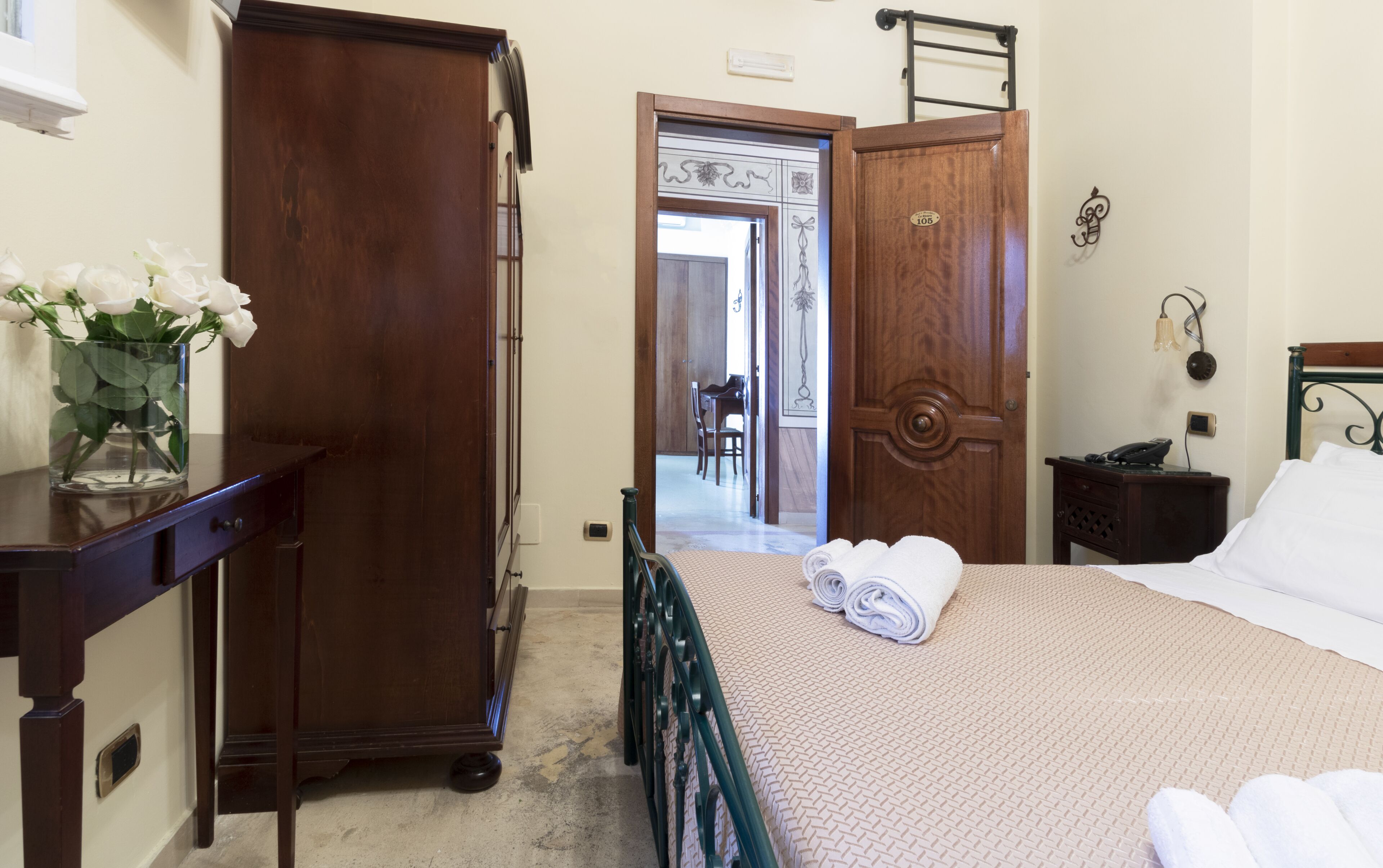A hotel room with a bed, a wooden wardrobe, a desk with a vase of flowers, and a bathroom visible through an open door.