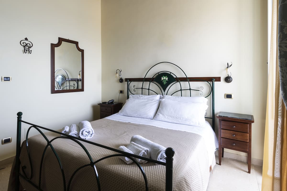 Un lit bien fait, avec du linge de lit blanc et des serviettes pliées, une petite table de chevet, un miroir fixé au mur et une commode.