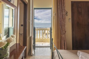 A room with a bed, a bedside table with a vase of flowers, a view of the sea through an open door, and a wooden door.