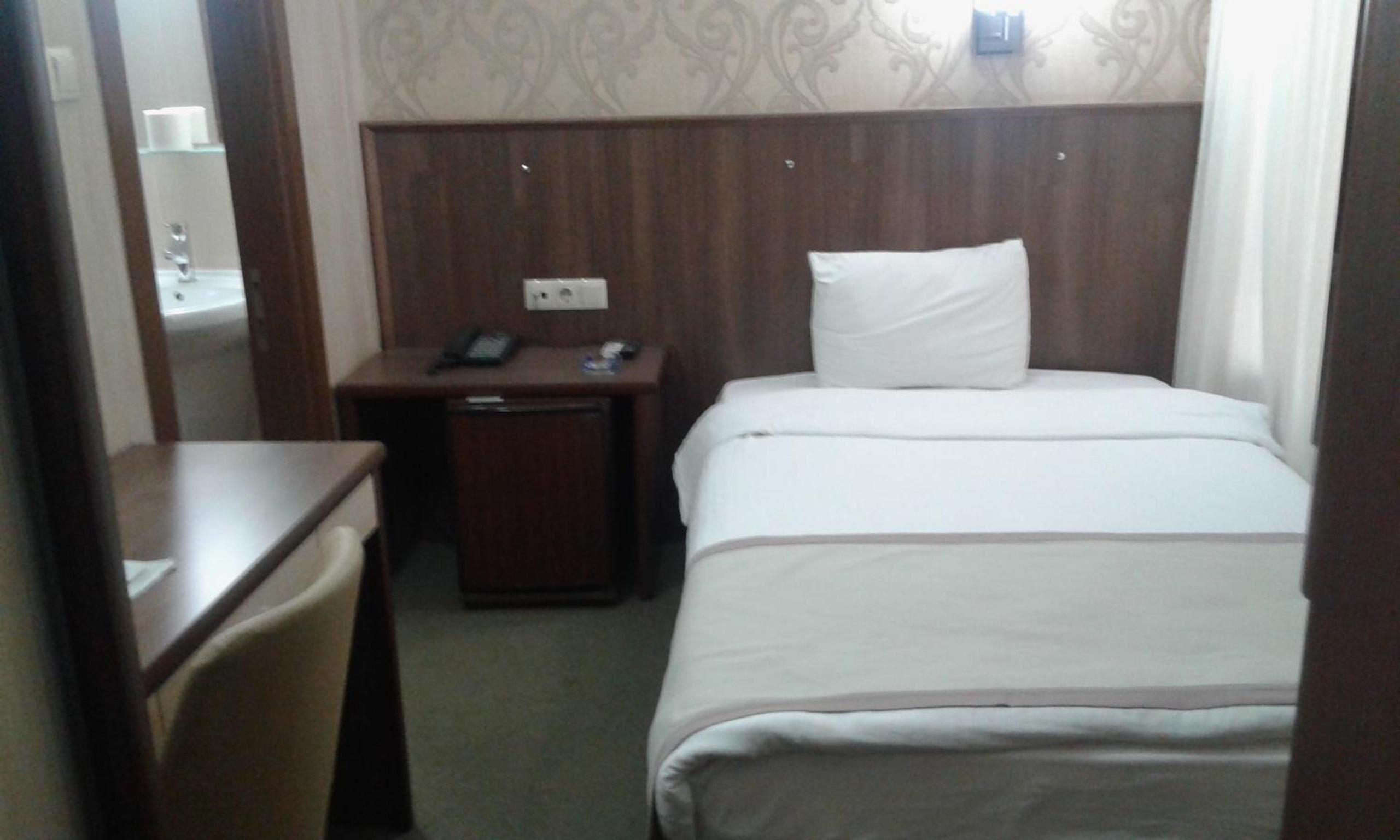 A hotel room with a bed, a bedside table, a chair, and a small desk with a phone and a lamp.