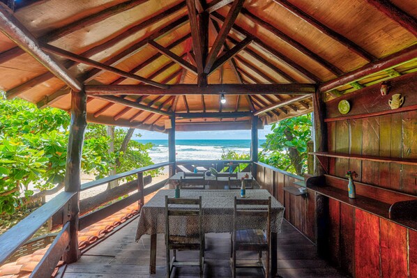 A covered outdoor seating area with a view of the ocean, wooden tables and chairs, and a rustic wooden wall with a shelf. - Pousada Terral Surf (Itacaré)