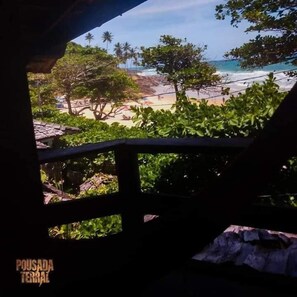 A beach view from a covered area with a wooden railing, palm trees, and a clear sky.