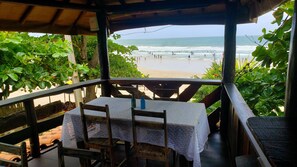 A covered outdoor seating area with a table and chairs, overlooking a beach with people and waves. - Pousada Terral Surf (Itacaré)