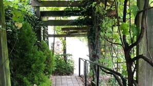 Ein gepflasterter Weg unter einer Holzpergola mit Metallgeländer, umgeben von Grünpflanzen und Bäumen.