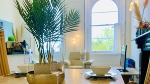 Ein Essbereich mit einem großen Tisch, der für eine Mahlzeit gedeckt ist, einer Topfpflanze und Blick ins Freie durch ein Fenster.
