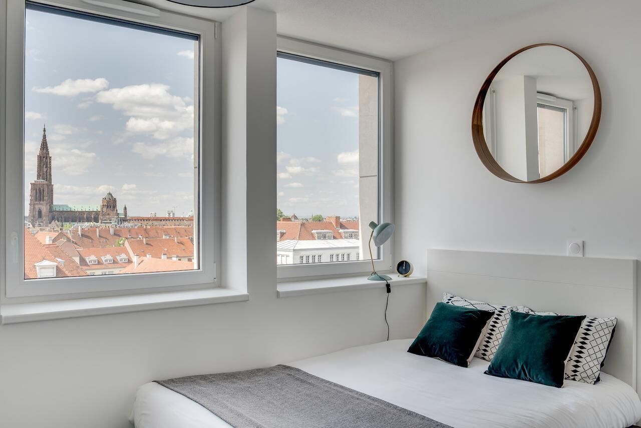 A bedroom with a large bed, two windows, a round mirror, and a view of a cityscape.