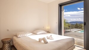 Quarto com cama, almofadas, mesa de cabeceira e vista para uma piscina e montanhas através de uma porta de vidro.