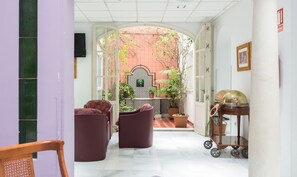 Un couloir donnant sur une cour intérieure, avec un mur recouvert de carreaux formant un motif géométrique, une plante en pot et un chariot en bois.