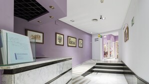 A hotel reception area with a marble counter, framed artwork on the walls, and a staircase leading to another section. - Hotel Platería by Bossh Hotels (Ecija)