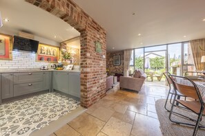 A kitchen with grey cabinets, a brick wall, and a tiled floor. There is a dining area with a table and chairs, and a view of the outdoors through large windows.
