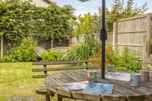 A wooden table with magazines and mugs, surrounded by a garden with a wooden fence and greenery. - Cog Cottage, Great Walsingham. Norfolk (Great Walsingham)