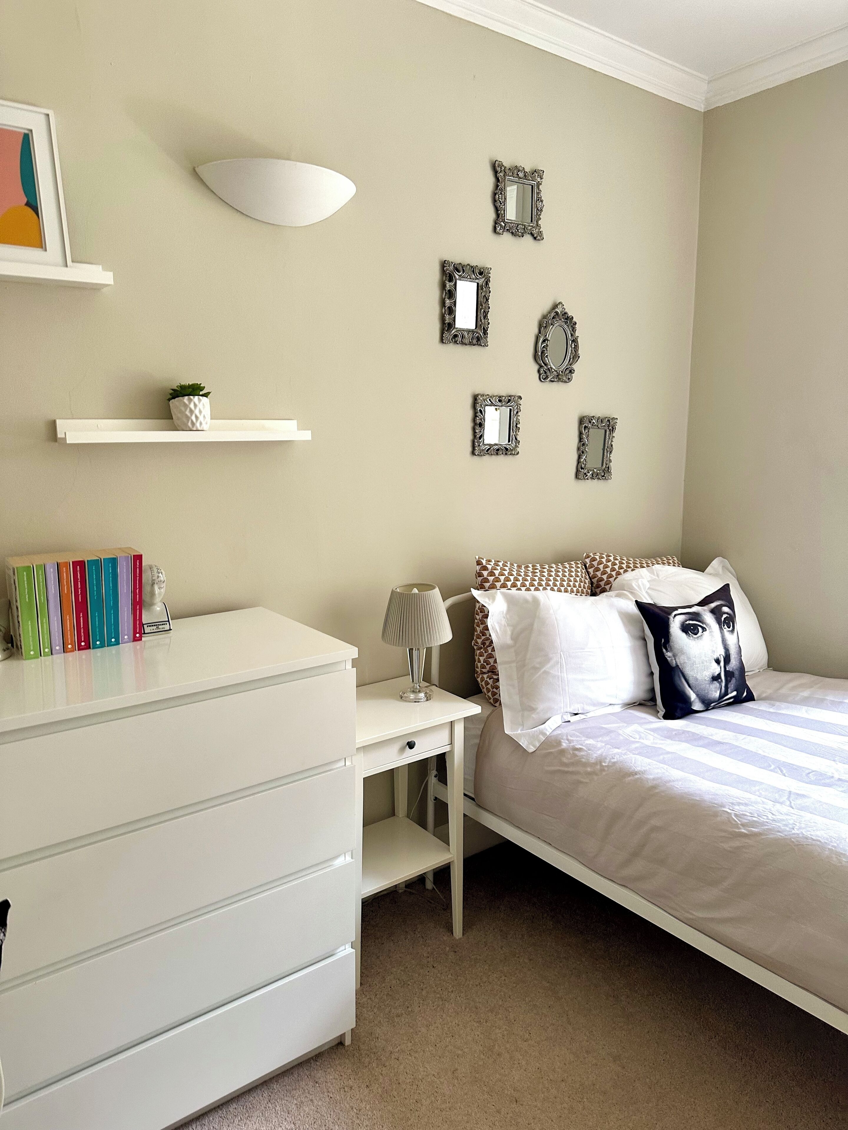 Une chambre avec une commode blanche, une table de chevet avec une lampe, un lit agrémenté d’oreillers à motifs et des miroirs fixés au mur.